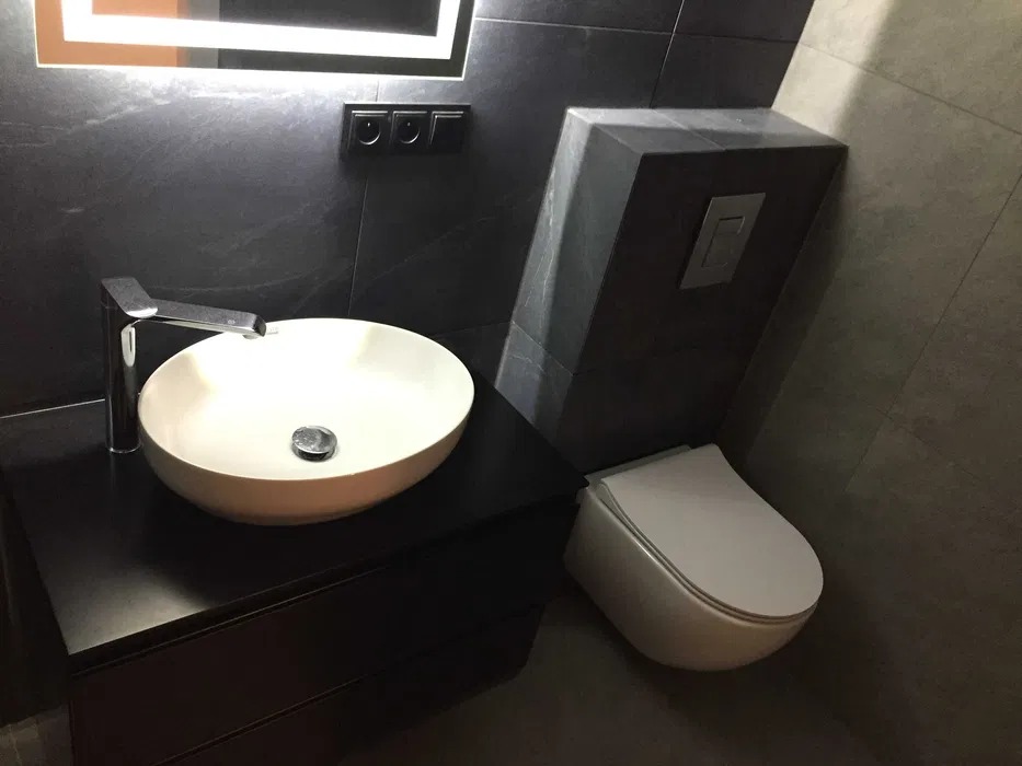 Nowoczesna łazienka w ciemnej tonacji z owalną umywalką nablatową, chromowaną baterią, podświetlanym lustrem i podtynkową toaletą.