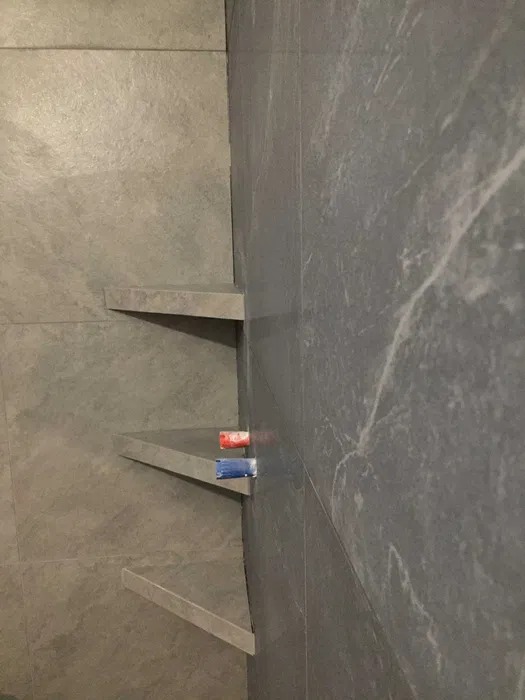 Wykończona ściana prysznicowa z szarych płytek imitujących kamień, z wbudowanymi półkami narożnymi z tego samego materiału. Na jednej z półek widoczne dwie zakrętki od kosmetyków w kolorach...