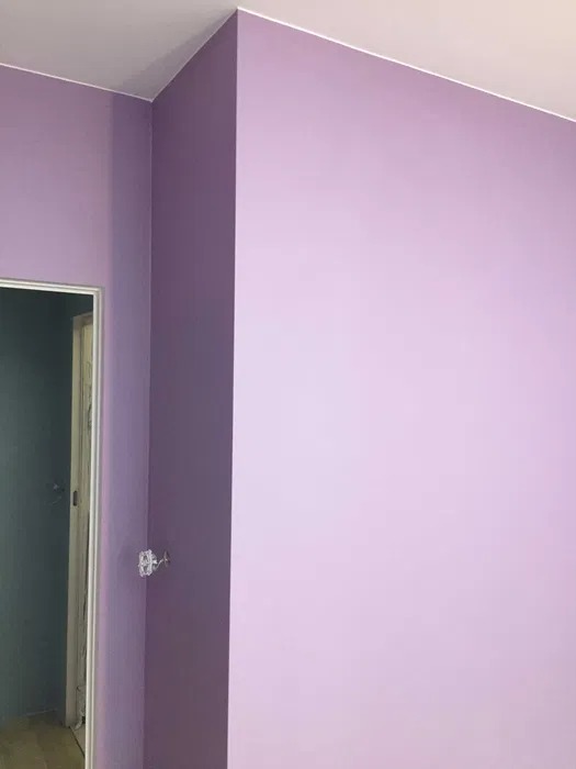Świeżo pomalowany róg pokoju w odcieniach fioletu, widoczne gniazdko elektryczne wystające ze ściany, drzwi w tle.
