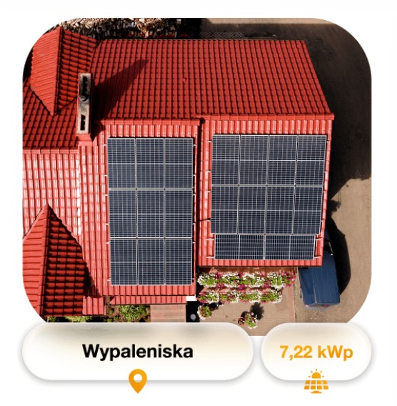 Widok z góry na czerwony dach domu z zainstalowanymi panelami fotowoltaicznymi; kwiaty w donicach pod panelami, samochód w tle.