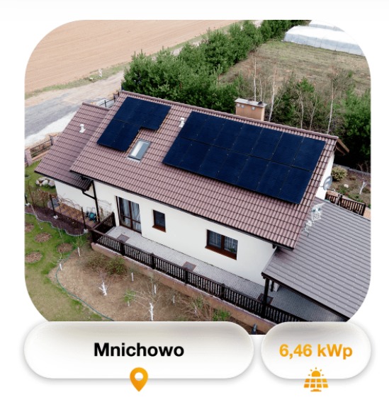 Widok z góry na dom jednorodzinny z brązowym dachem, na którym zamontowano panele słoneczne; widoczne także drzewa i pole w tle.