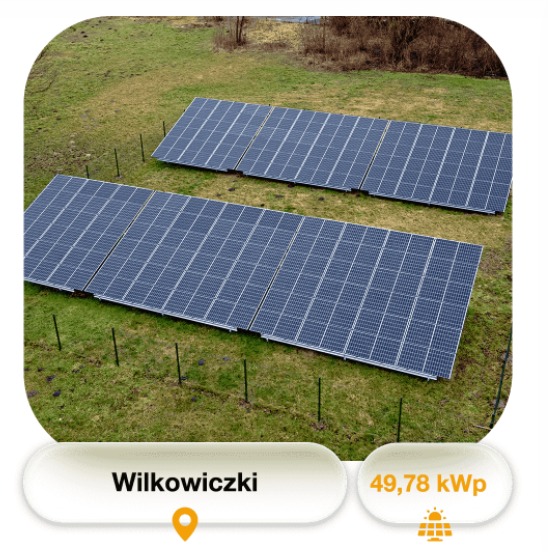 Dwa rzędy paneli słonecznych na zielonym, lekko pofałdowanym terenie, ogrodzone niskim płotem z czarnych słupków, widok z góry.