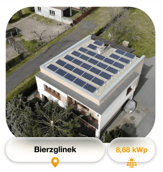 Widok z góry na dom z płaskim dachem pokrytym panelami słonecznymi ułożonymi w rzędach, otoczony zielenią i drogą.