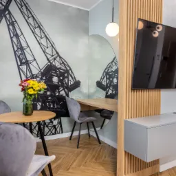 Wnętrze apartamentu w Gdańsku z fototapetą przedstawiającą dźwigi portowe, okrągłym lustrem nad biurkiem, drewnianymi lamelami na ścianie, telewizorem, szarą komodą, okrągłym stołem z bukietem...