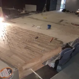 Układanie podłogi z płytek imitujących drewno w dużej przestrzeni, widoczne krzyżyki dystansowe, wiaderko z klejem Dekoral i częściowo zasłonięte krzesła kinowe w tle.