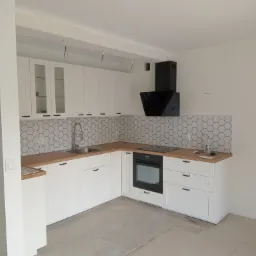 Jasna kuchnia z białymi szafkami, drewnianym blatem i heksagonalnymi płytkami na ścianie, czarny okap nad płytą grzewczą, widoczne kable zwisające z sufitu, podłoga częściowo zabezpieczona kartonem.