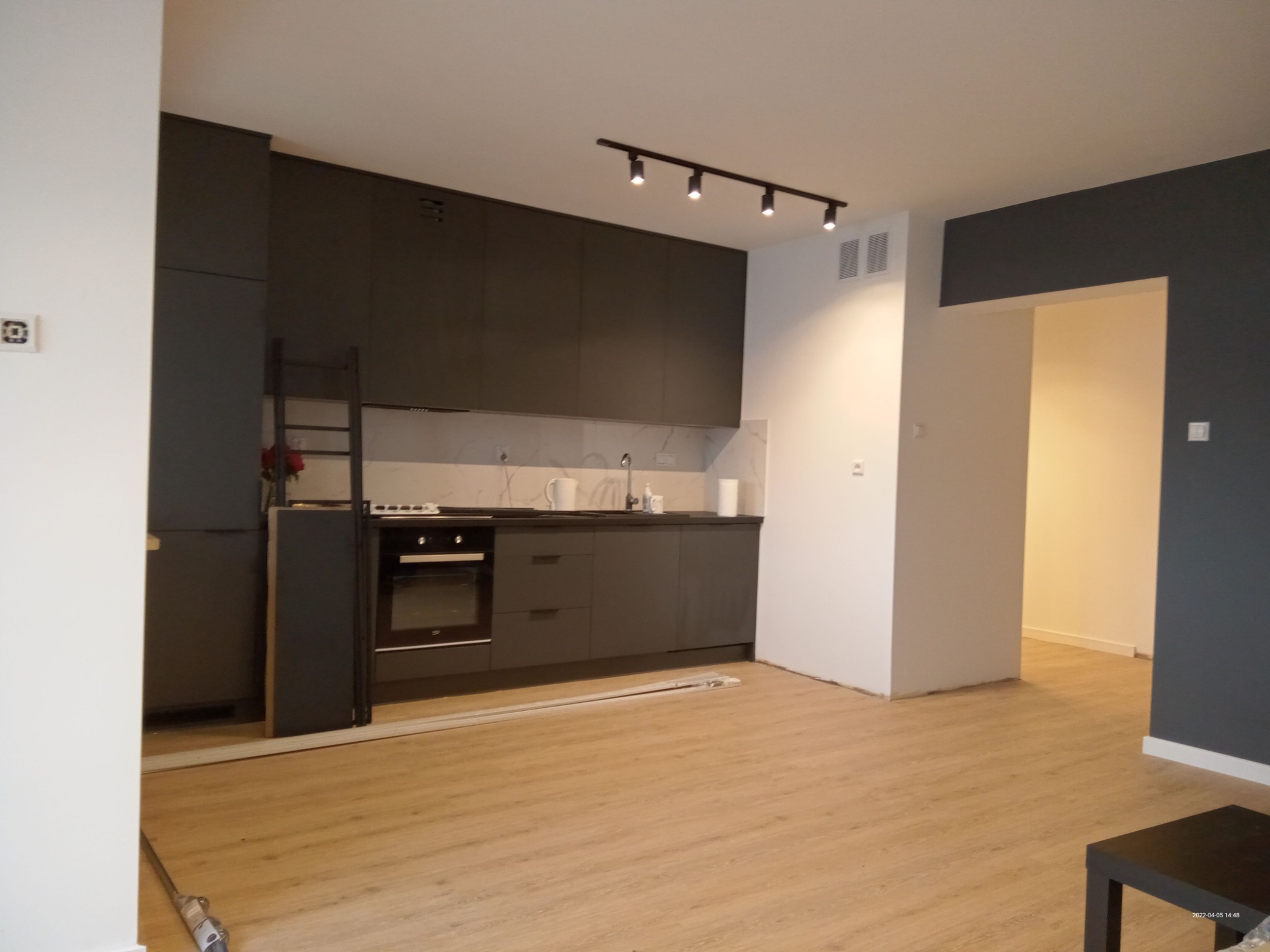 Nowoczesna kuchnia z ciemnymi, matowymi szafkami i marmurowym blatem, oświetlona listwą z reflektorami, z widocznymi listwami przypodłogowymi i fragmentem podłogi w trakcie układania paneli.