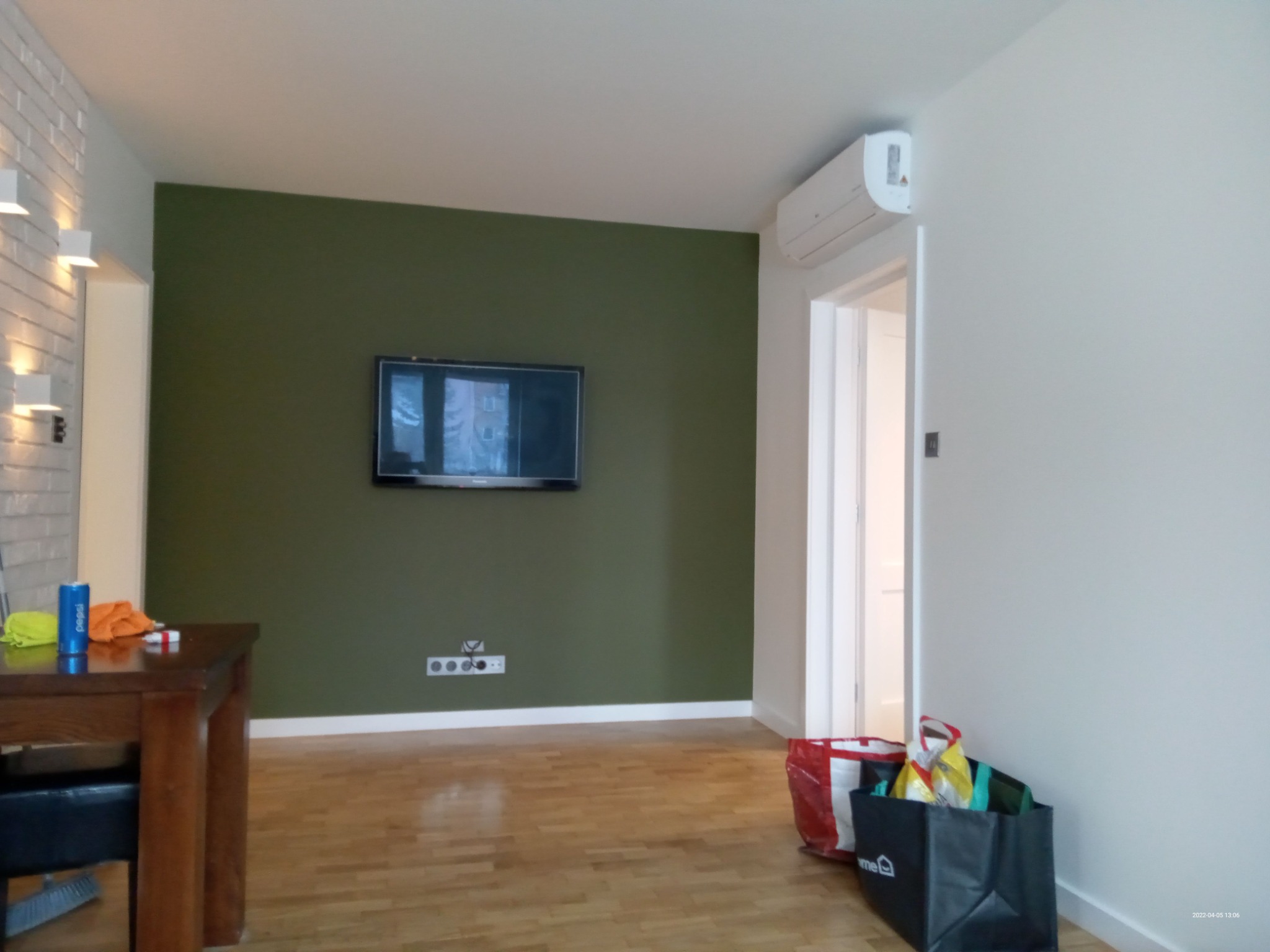 Wnętrze pokoju z zieloną ścianą, telewizorem, klimatyzacją i drewnianą podłogą, w trakcie remontu lub wykończenia, widoczne torby z zakupami i niebieska puszka na stole.