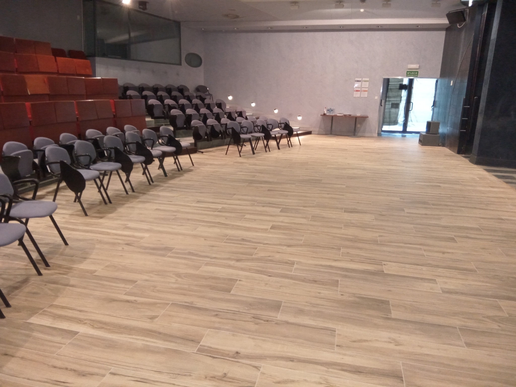 Sala konferencyjna z rzędami krzeseł i podłogą wyłożoną panelami imitującymi drewno, widok na scenę i wyjście ewakuacyjne.