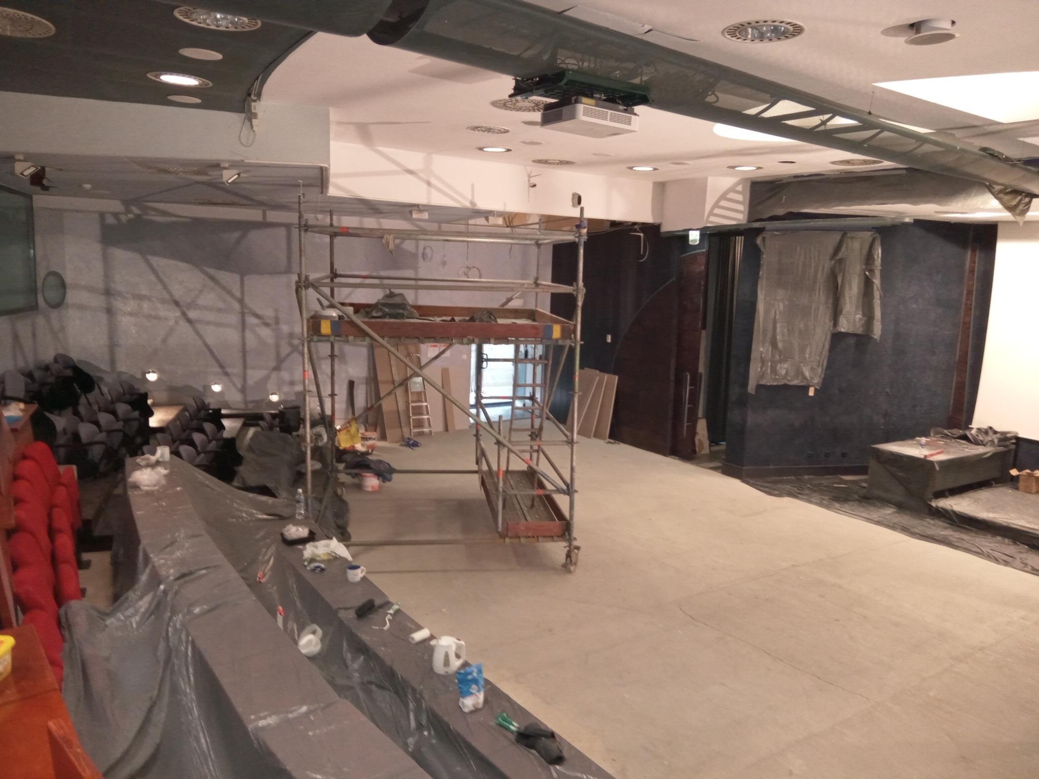 Sala w trakcie remontu: rusztowanie, zakryte folią krzesła, narzędzia malarskie i materiały budowlane. Widoczny projektor pod sufitem i okrągłe lampy.