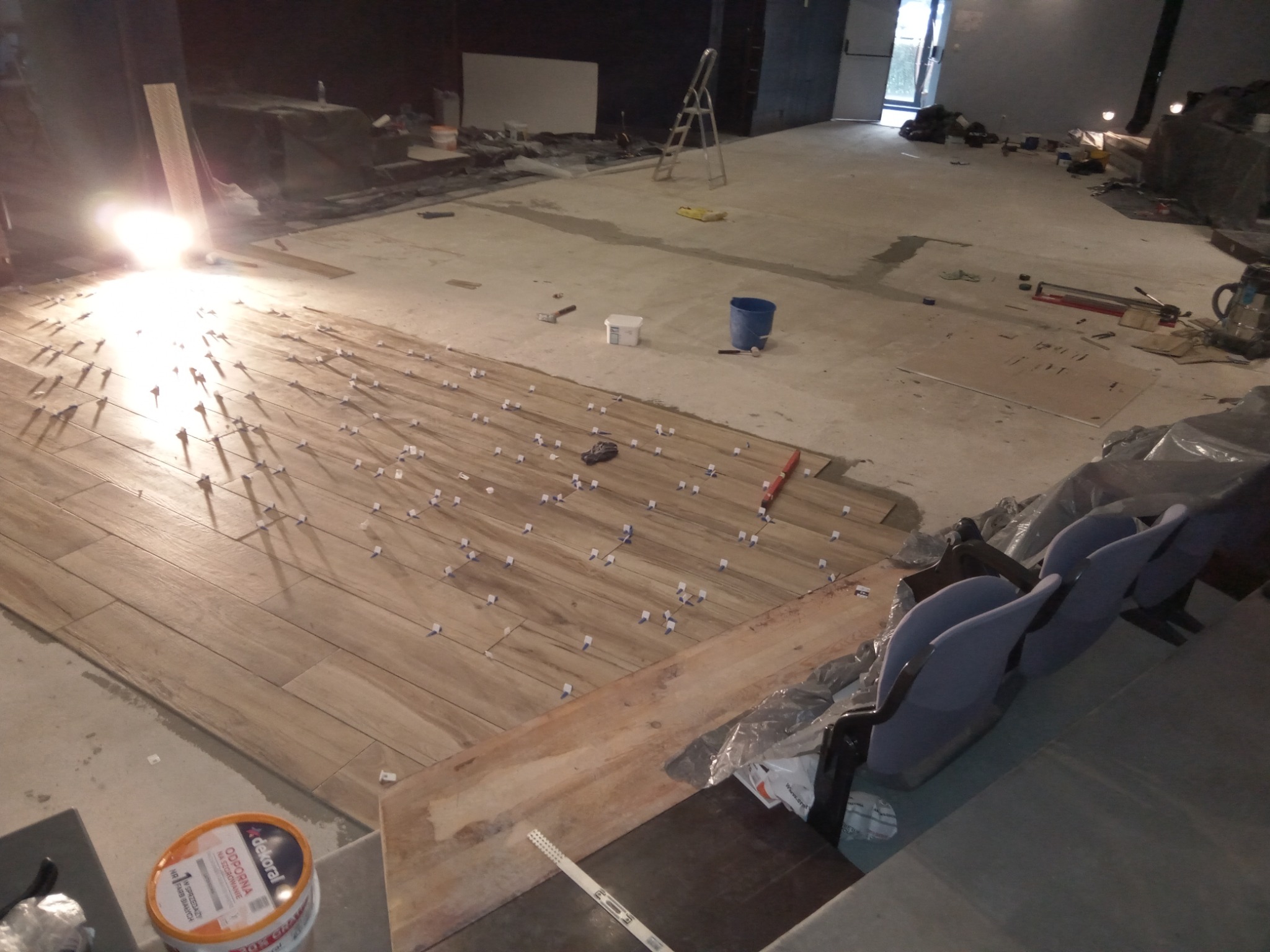 Układanie podłogi z płytek imitujących drewno w dużej przestrzeni, widoczne krzyżyki dystansowe, wiaderko z klejem Dekoral i częściowo zasłonięte krzesła kinowe w tle.