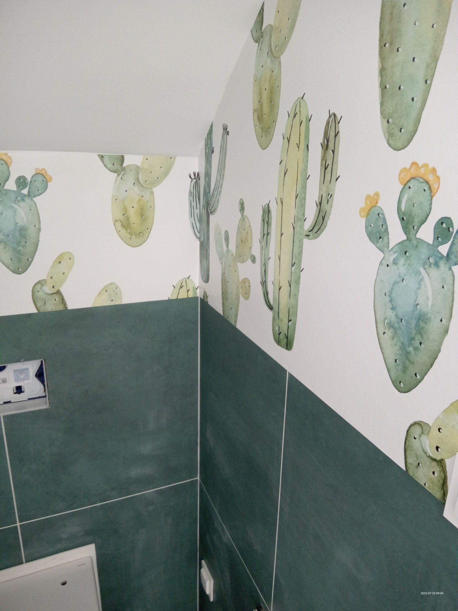 Łazienka z zielonymi płytkami i ścianą ozdobioną motywem akwarelowych kaktusów, widoczny fragment zabudowy WC.