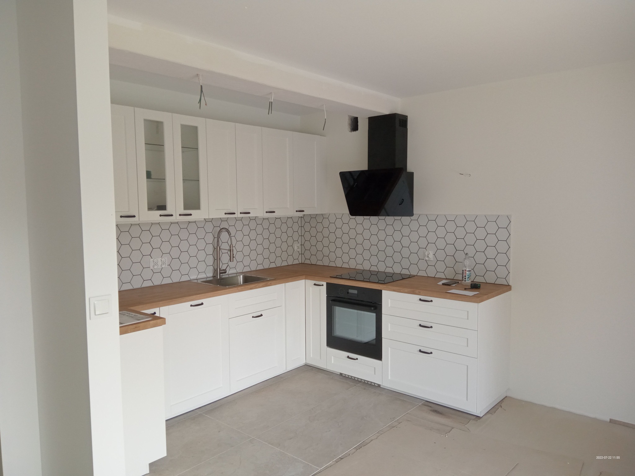 Jasna kuchnia z białymi szafkami, drewnianym blatem i heksagonalnymi płytkami na ścianie, czarny okap nad płytą grzewczą, widoczne kable zwisające z sufitu, podłoga częściowo zabezpieczona kartonem.