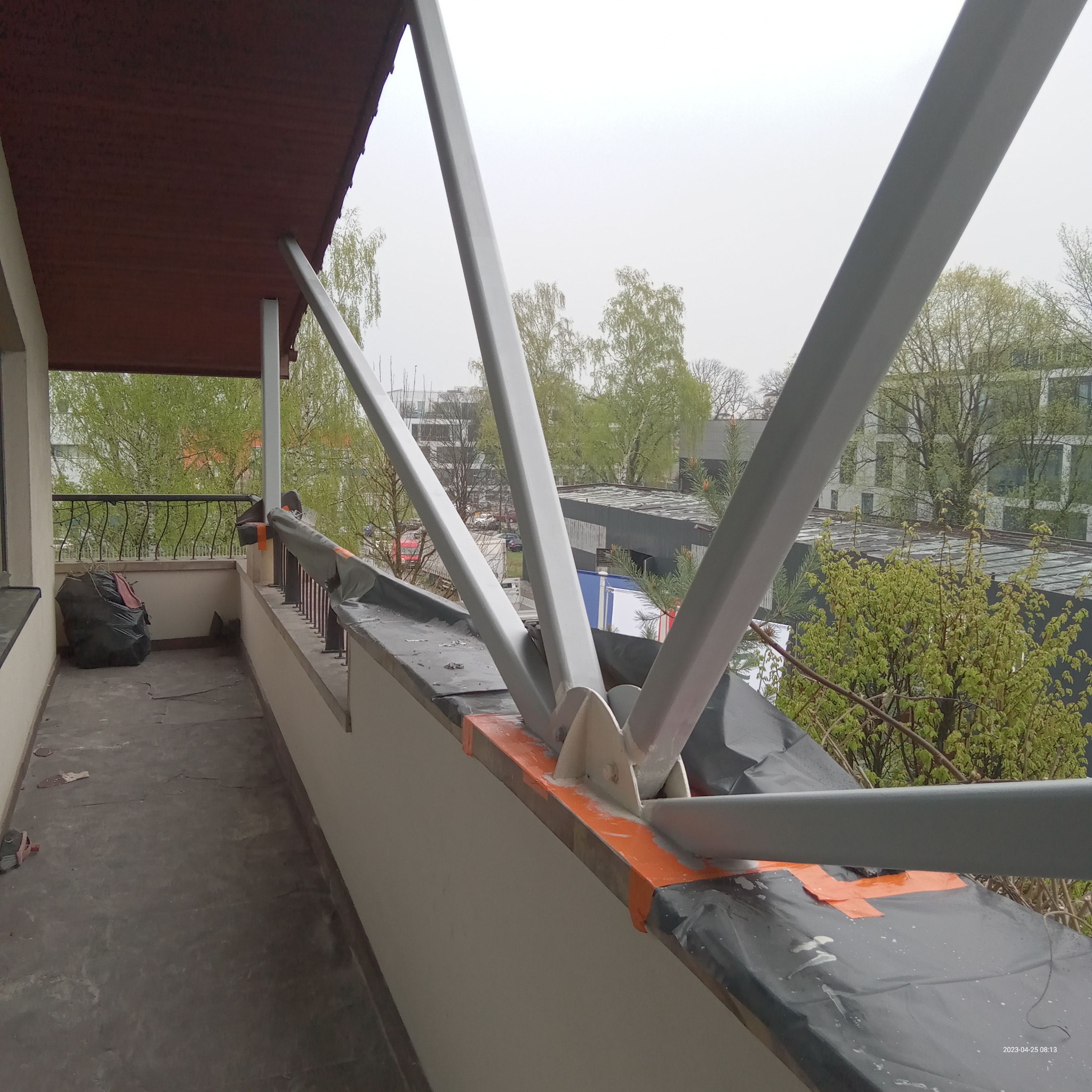 Montaż konstrukcji wsporczej z metalowych profili na balkonie, zabezpieczona folią i taśmą malarską, widok na zewnątrz z drzewami i budynkami w tle.