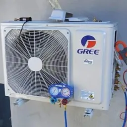 Biała jednostka zewnętrzna klimatyzacji GREE zamontowana na ścianie, podłączona do manometr&oacute;w i miernika cęgowego podczas diagnostyki lub serwisu.