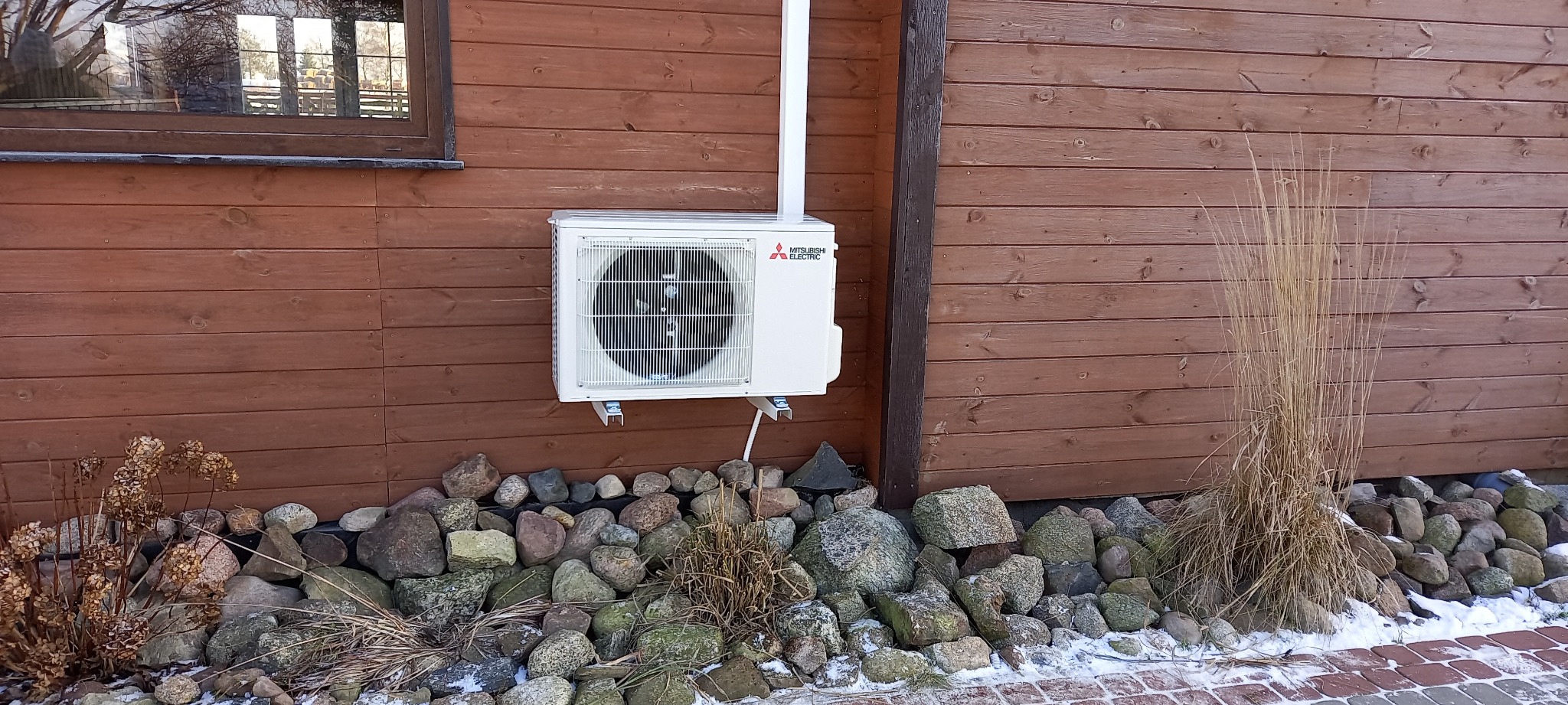 Zewnętrzna jednostka klimatyzacji Mitsubishi Electric zamontowana na drewnianej ścianie domu, z widocznymi elementami instalacji i kamieniami u podstawy.