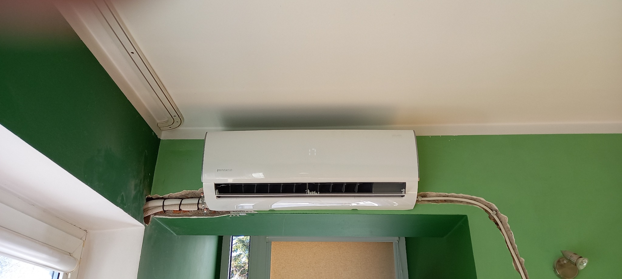 Biały klimatyzator Rotenso na zielonej ścianie, widoczne kable w korytach i niedokończone obróbki ściany po montażu.