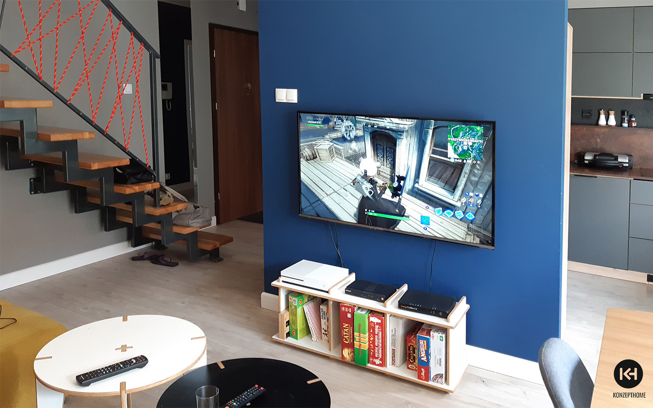 Nowoczesny salon z niebieską ścianą, telewizorem z grą wideo, drewnianymi schodami z czerwoną liną i niskim regałem z grami planszowymi.