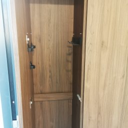 Ewkon - Wnętrze szafy z drewnianymi panelami, wyposażone w czarne uchwyty i wieszaki. Widoczne zawiasy i elementy mocujące. Minimalistyczny design.