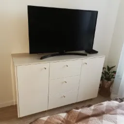 Jasny, lakierowany kredens z trzema szufladami i złotymi uchwytami, na którym stoi telewizor. Obok stoi roślina w wiklinowym koszu.