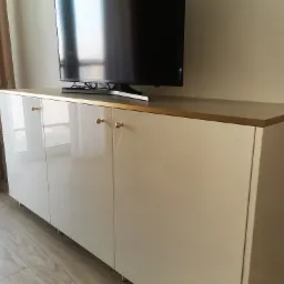 Biała, lakierowana szafka RTV z zaokrąglonym frontem, złotymi uchwytami i blatem, ustawiona na drewnianej podłodze pod telewizorem. Nowoczesny design.