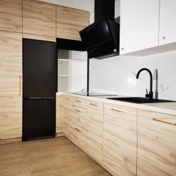 Ewkon - Nowoczesna kuchnia na wymiar z drewnianymi frontami, czarnym sprzętem AGD i złotymi uchwytami, wykończona białymi szafkami i marmurową ścianą.