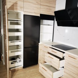 Ewkon - Jasna kuchnia z drewnopodobnymi frontami: lodówka w zabudowie, szafka cargo z wysuwanymi półkami, otwarte szuflady z podłużnymi uchwytami.
