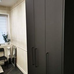 Ewkon - Ciemnoszara szafa trzydrzwiowa z podłużnymi uchwytami w łazience z czarną marmurową podłogą i jasnymi płytkami ściennymi. Widoczny fragment rośliny i akcesoriów łazienkowych.