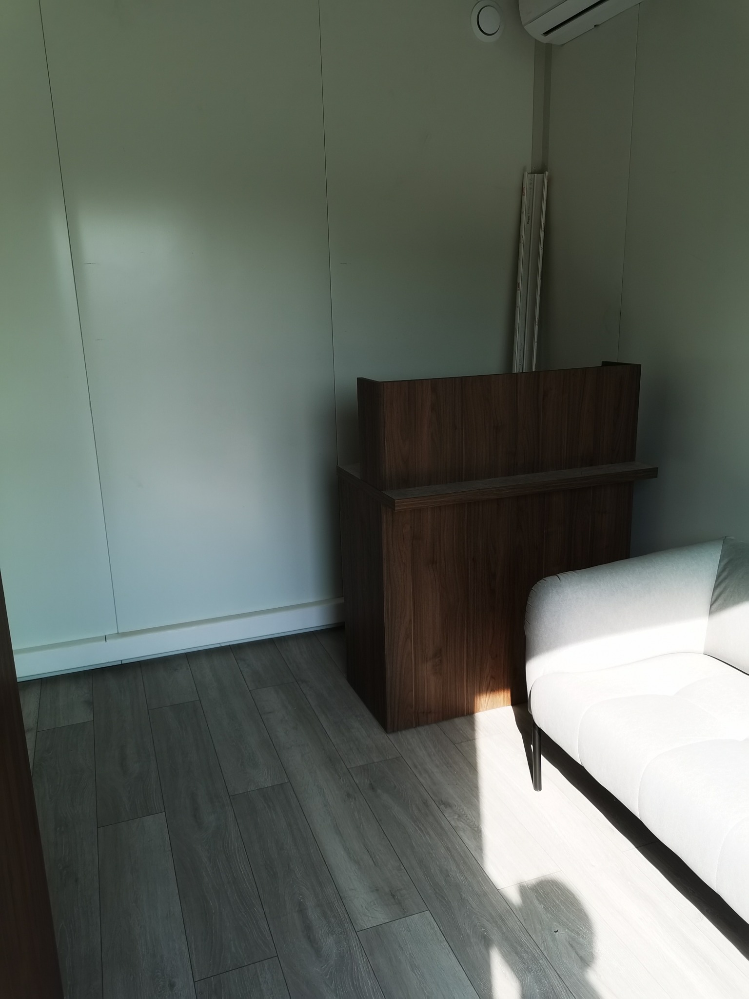 Nowoczesne wnętrze z drewnianą zabudową i białą sofą, oświetlone naturalnym światłem. Minimalistyczny design z akcentem na detale wykończeniowe i parkiet w odcieniach szarości.