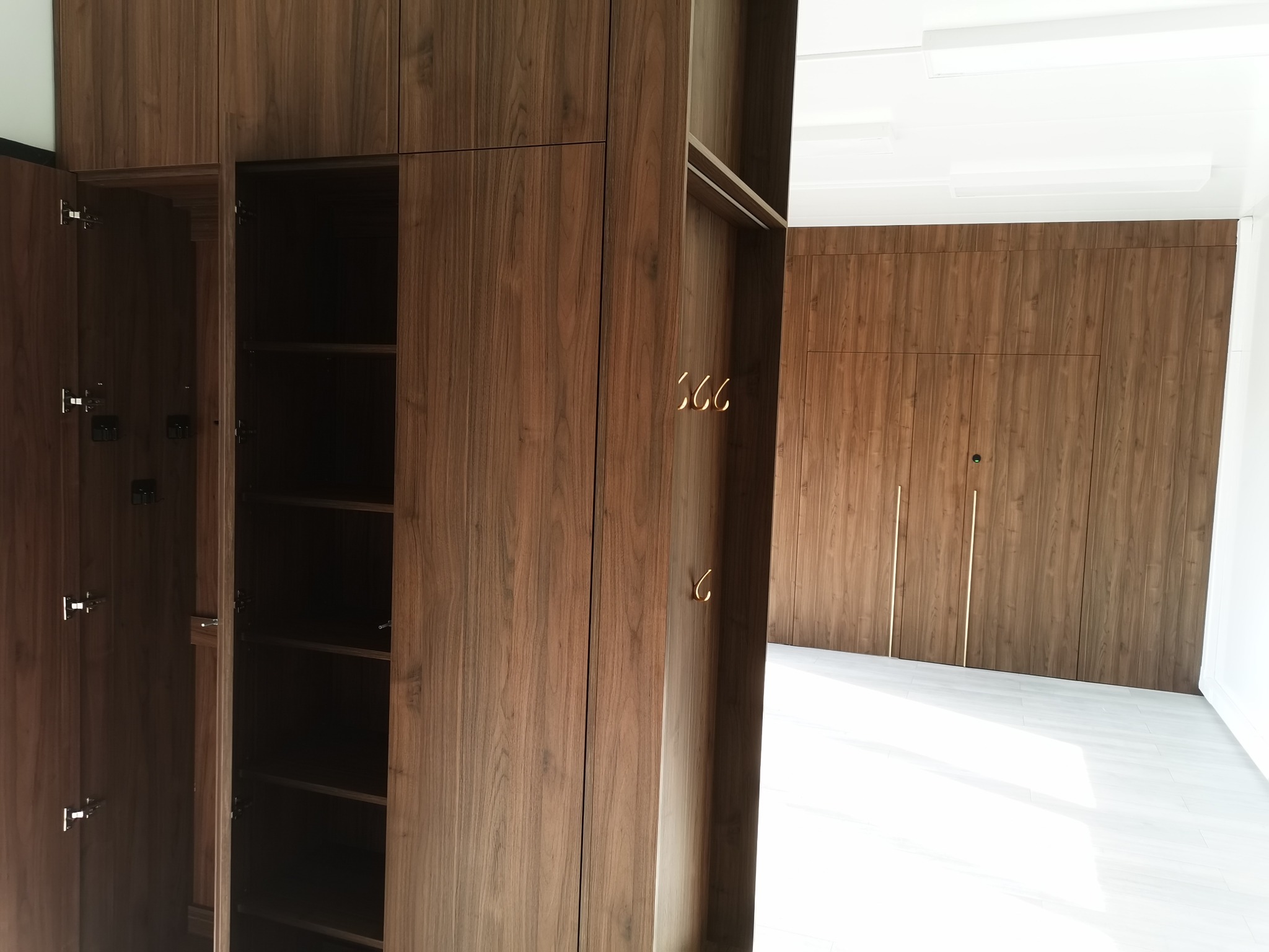 Wnętrze z zabudową z forniru orzechowego: szafa z otwartymi drzwiami, widoczne półki. Na ścianie w głębi wbudowane szafy z pionowymi, złotymi uchwytami.