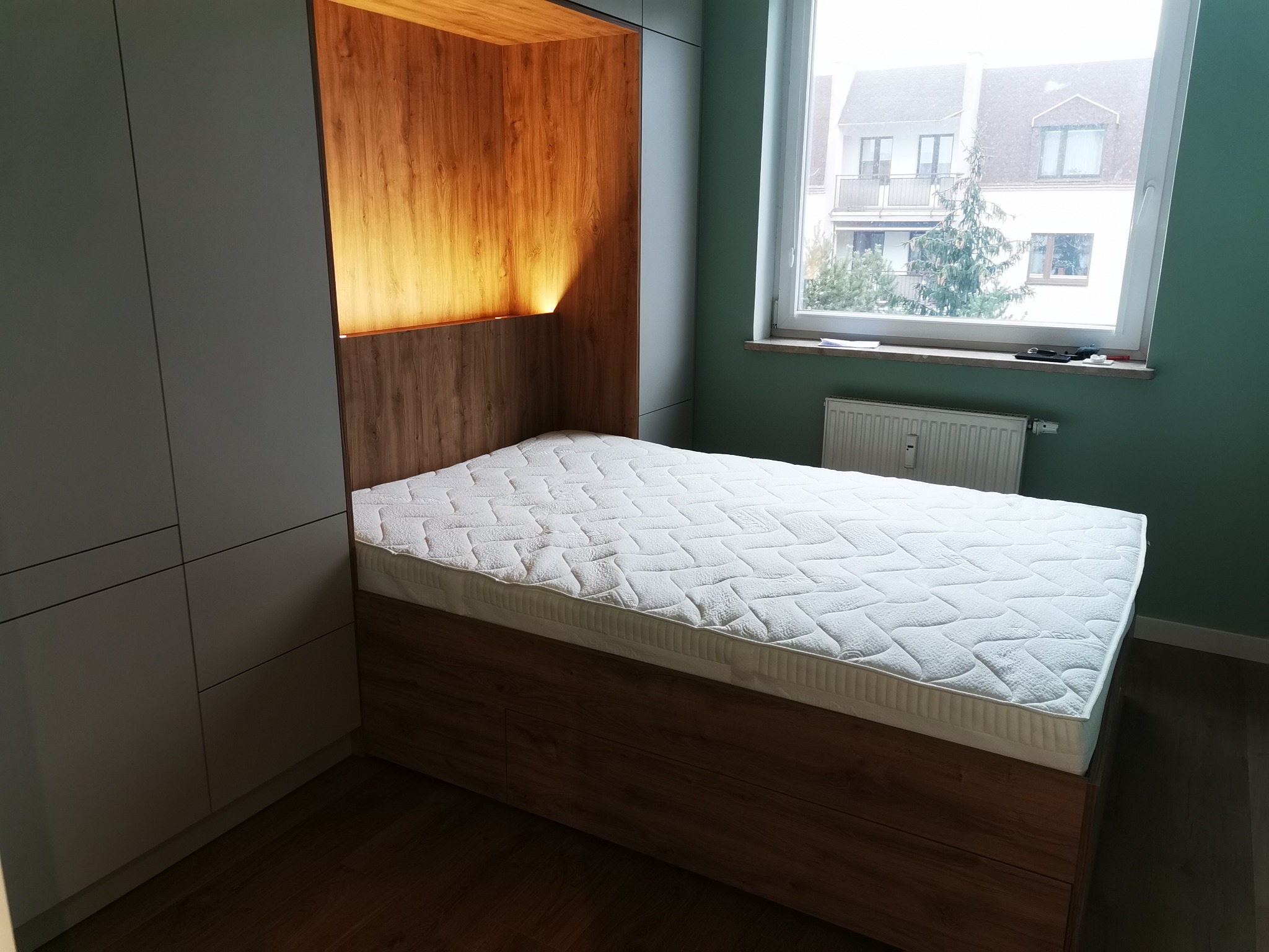 Nowoczesna szafa wnękowa z drewnianym wykończeniem i wbudowanym łóżkiem, oświetlona od góry, na tle okna z widokiem na budynek.