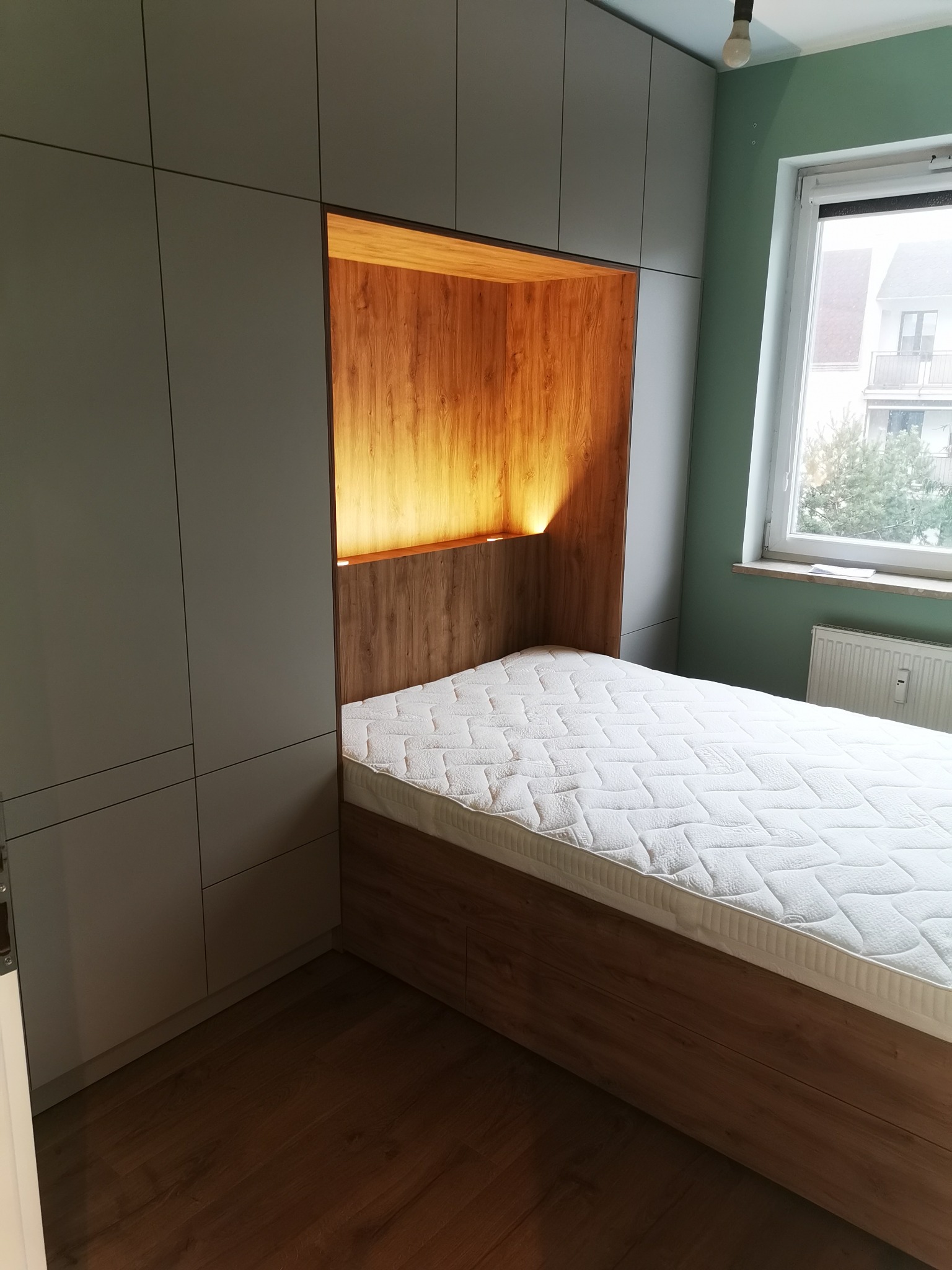 Szara szafa wnękowa z drewnianą wnęką na łóżko i oświetleniem. W tle okno. Minimalistyczny design szafy z subtelnymi uchwytami. Widok na materac.