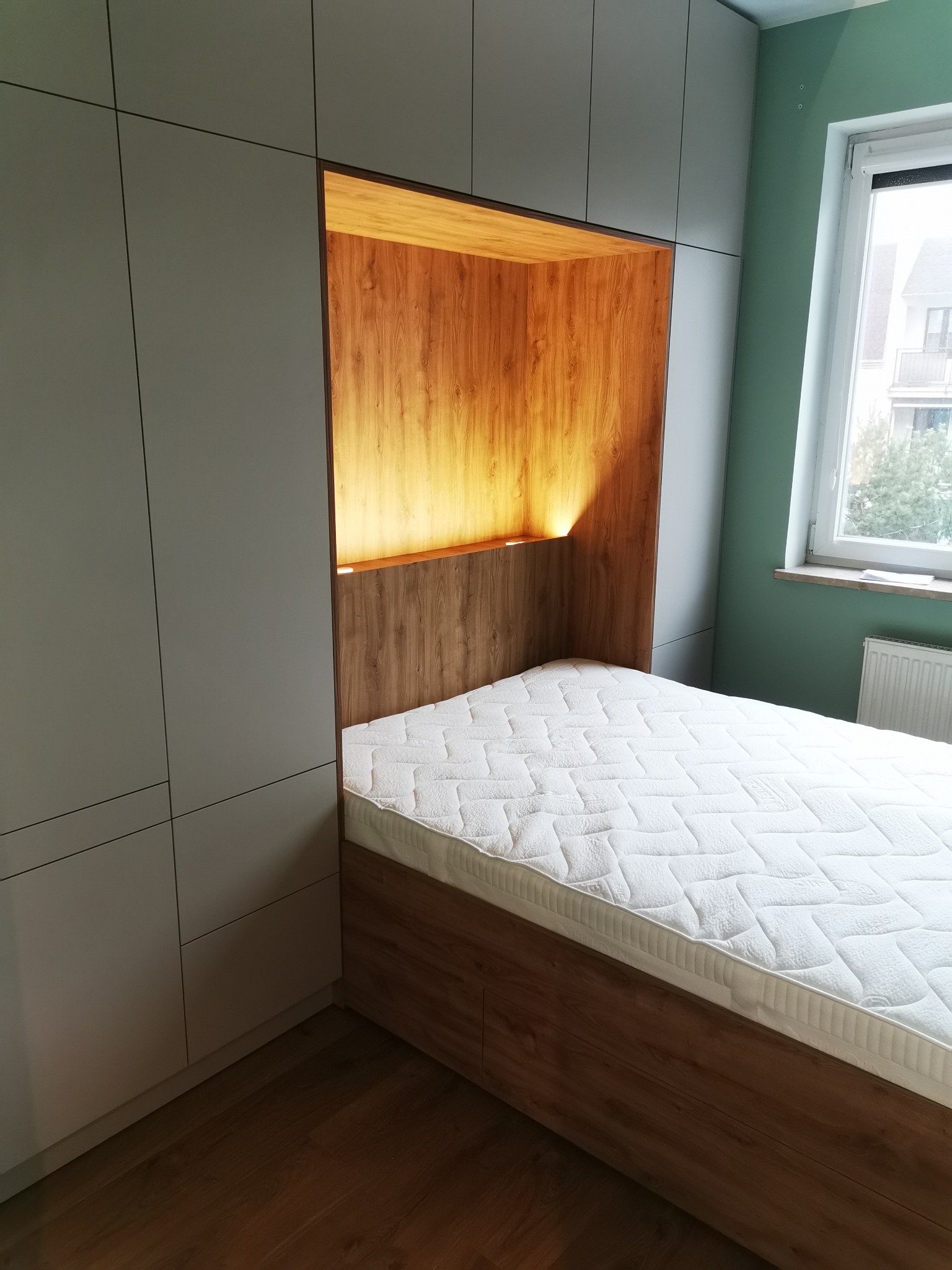 Szara szafa wnękowa z drewnianą wnęką nad łóżkiem. Wnęka podświetlona, widoczny materac. Minimalistyczny design, jasne wnętrze.