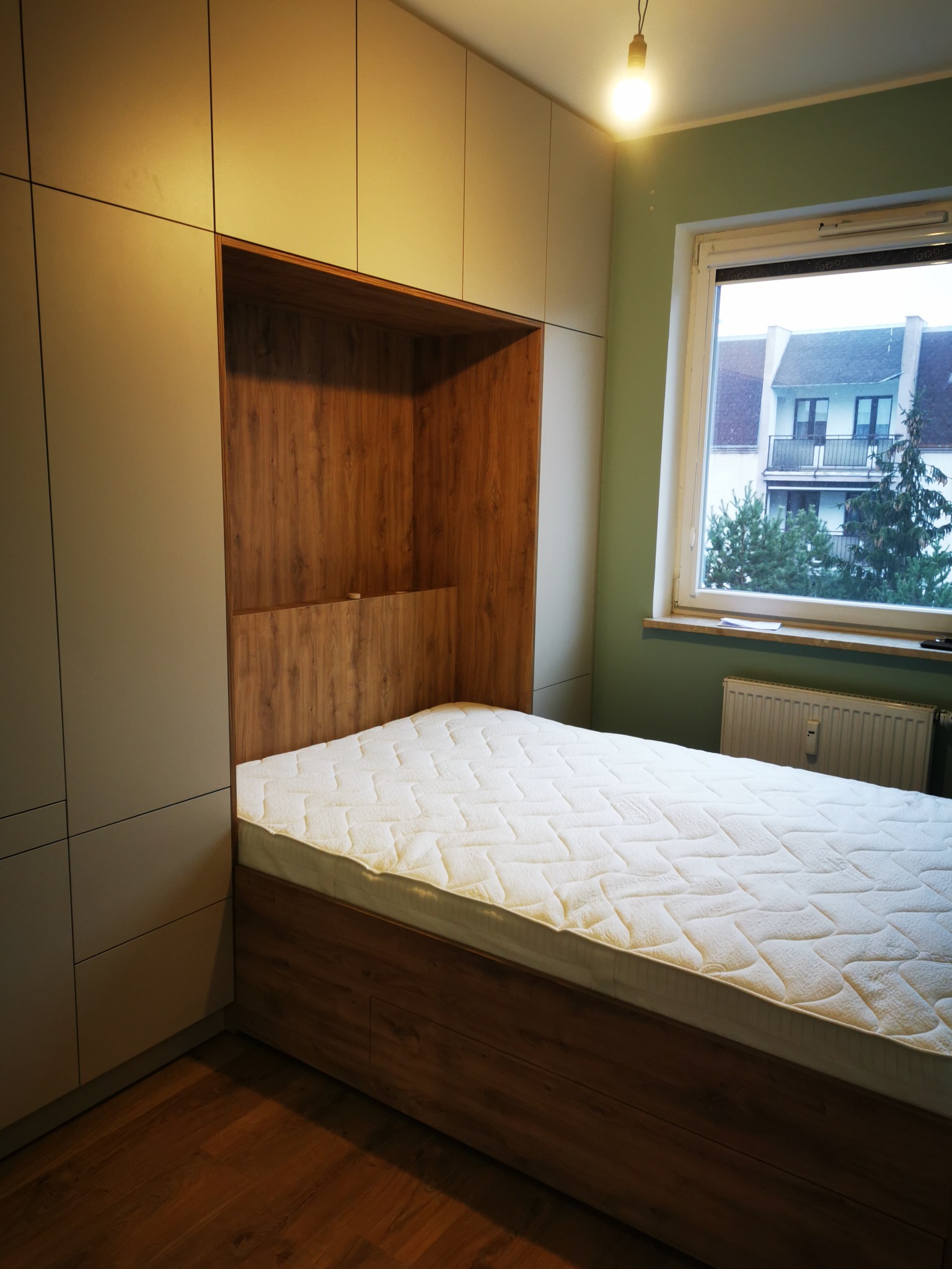 Szafa wnękowa z drewnianą wnęką na łóżko, materac z jasnym pokrowcem. U góry rząd szafek, z boku okno z widokiem na budynek.