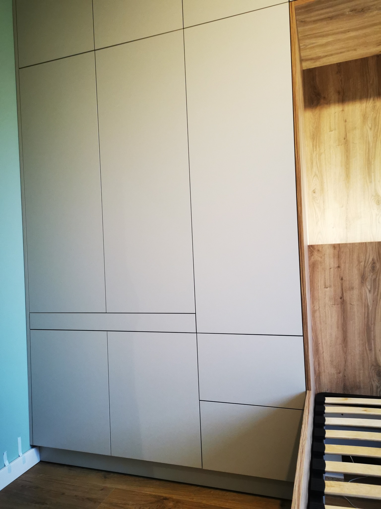 Szara szafa wnękowa z minimalistycznymi, ciemnymi liniami podziału frontów, przylegająca do drewnianego boku łóżka i turkusowej ściany.