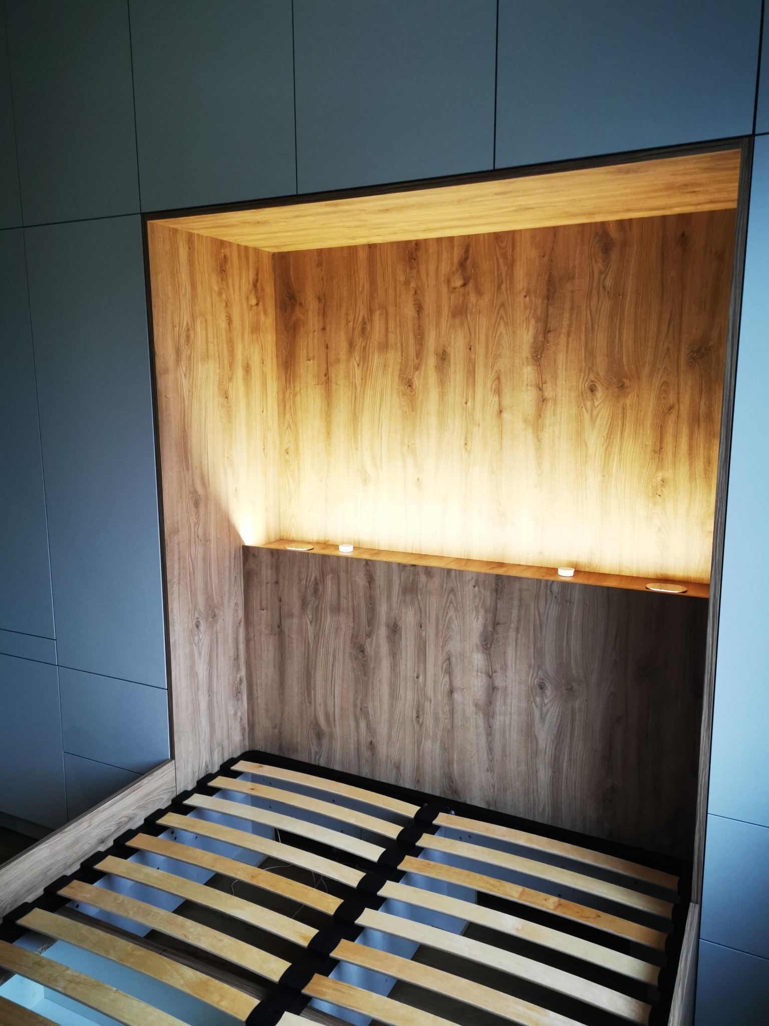 Narożna szafa wnękowa z drewnianą wnęką na łóżko, oświetloną od góry. Widoczny stelaż łóżka i fragmenty niebieskich frontów szafy.
