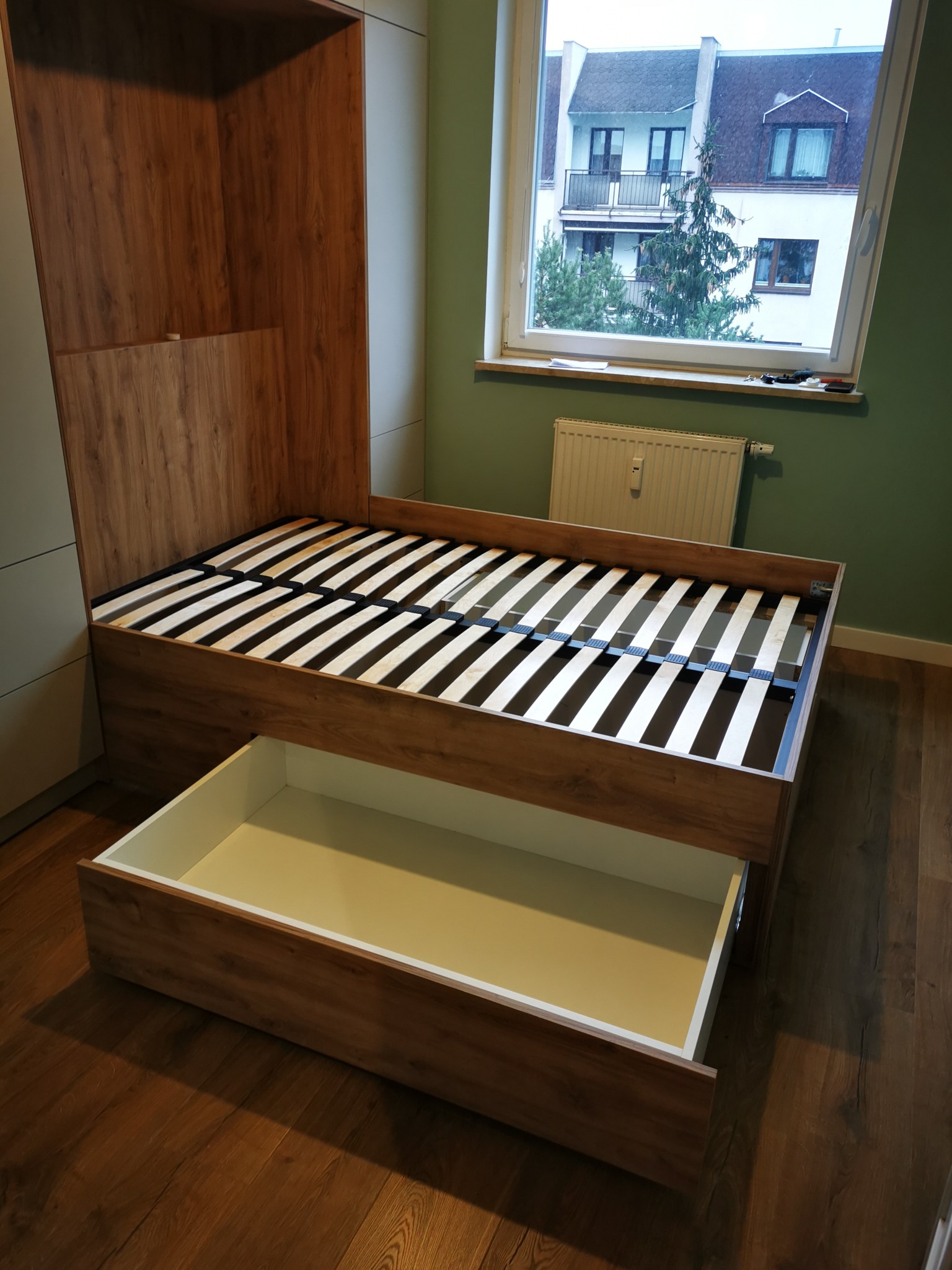 Rozkładane łóżko w szafie z drewnianym wykończeniem i wysuwaną szufladą, na tle okna z widokiem na budynek mieszkalny. Nowoczesne rozwiązanie do małych przestrzeni.