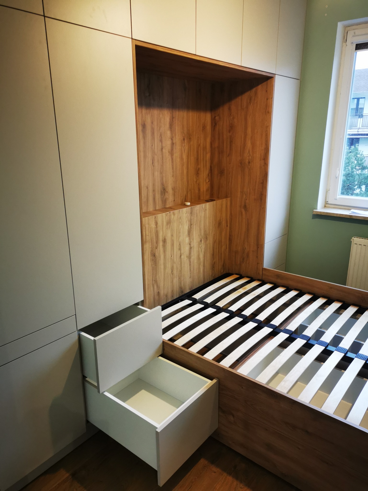 Szafa wnękowa z drewnianym zagłówkiem i wysuwanymi szufladami w jasnym pokoju, widok na stelaż łóżka i okno. Minimalistyczny design.