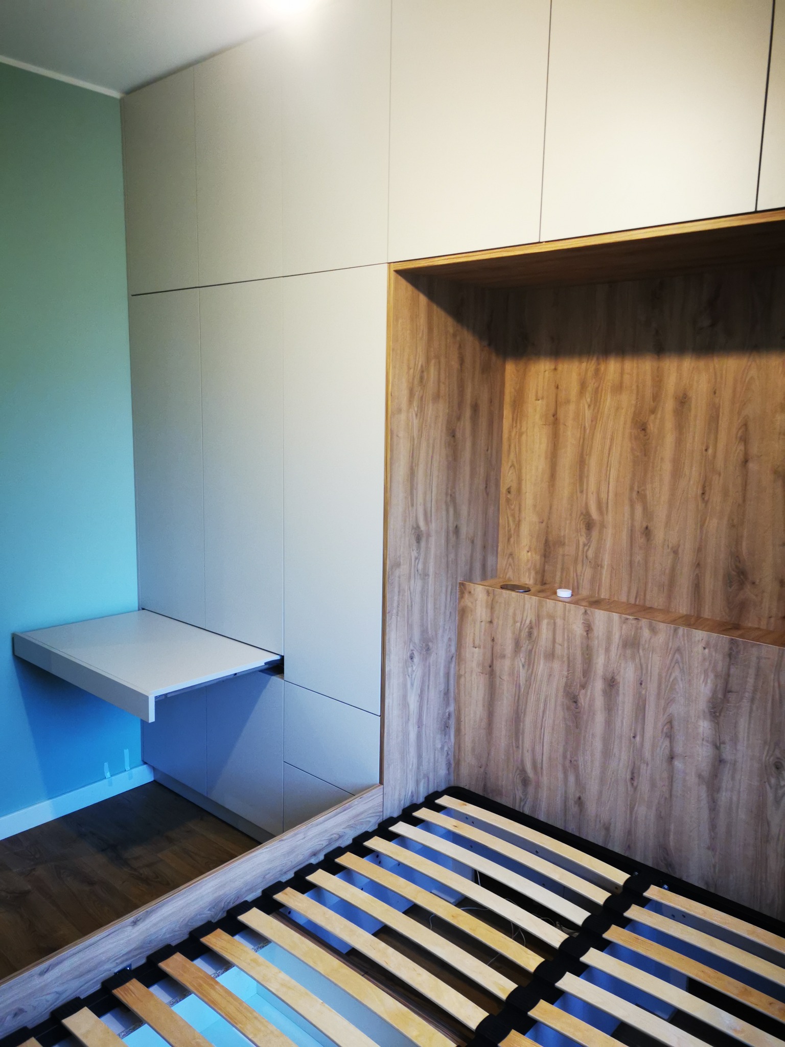 Minimalistyczna szafa wnękowa z drewnianym wykończeniem, wkomponowana w przestrzeń sypialni z łóżkiem i biurkiem. Jasna kolorystyka i proste linie.