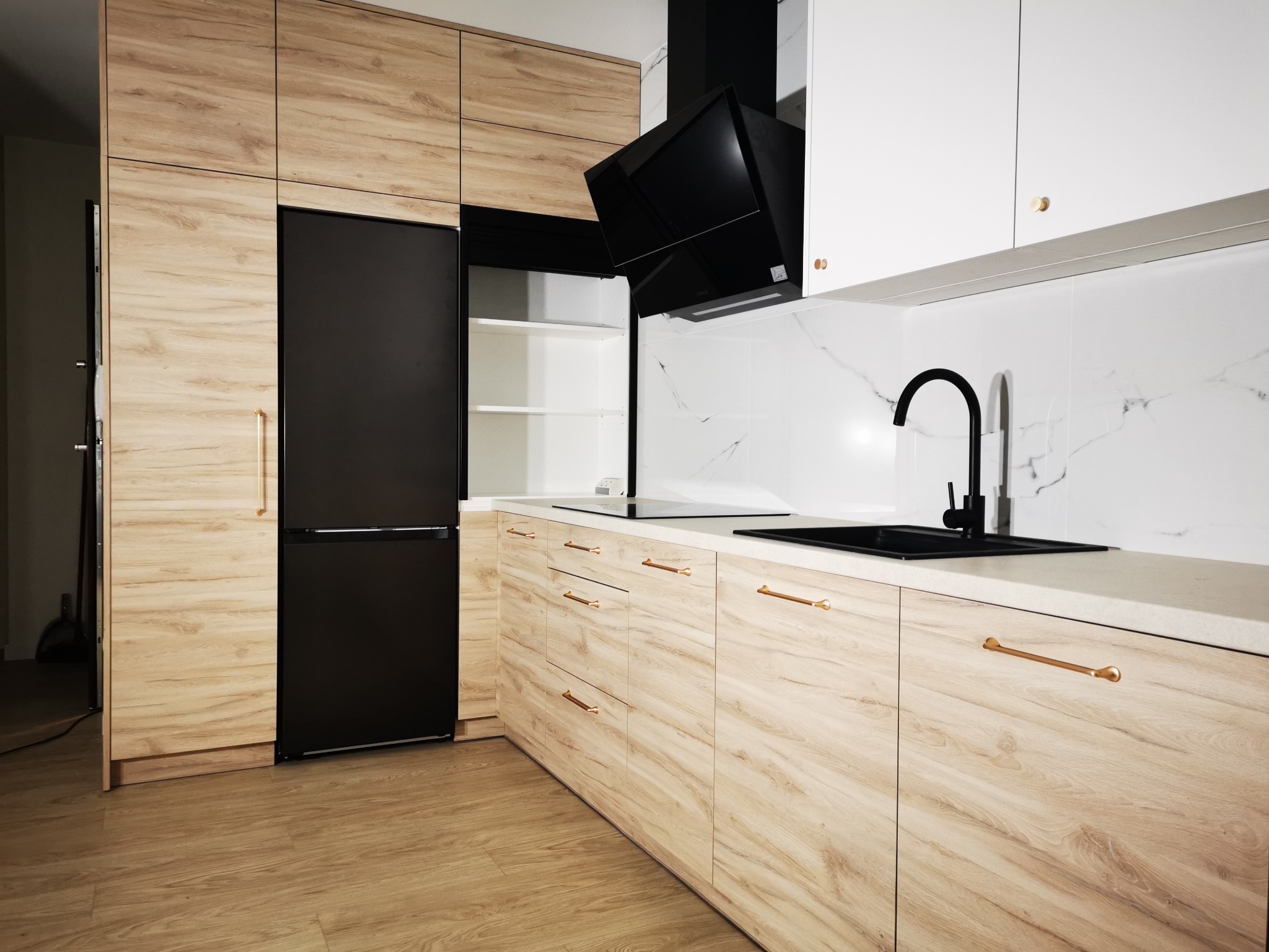 Nowoczesna kuchnia na wymiar z drewnianymi frontami, czarnym sprzętem AGD i złotymi uchwytami, wykończona białymi szafkami i marmurową ścianą.