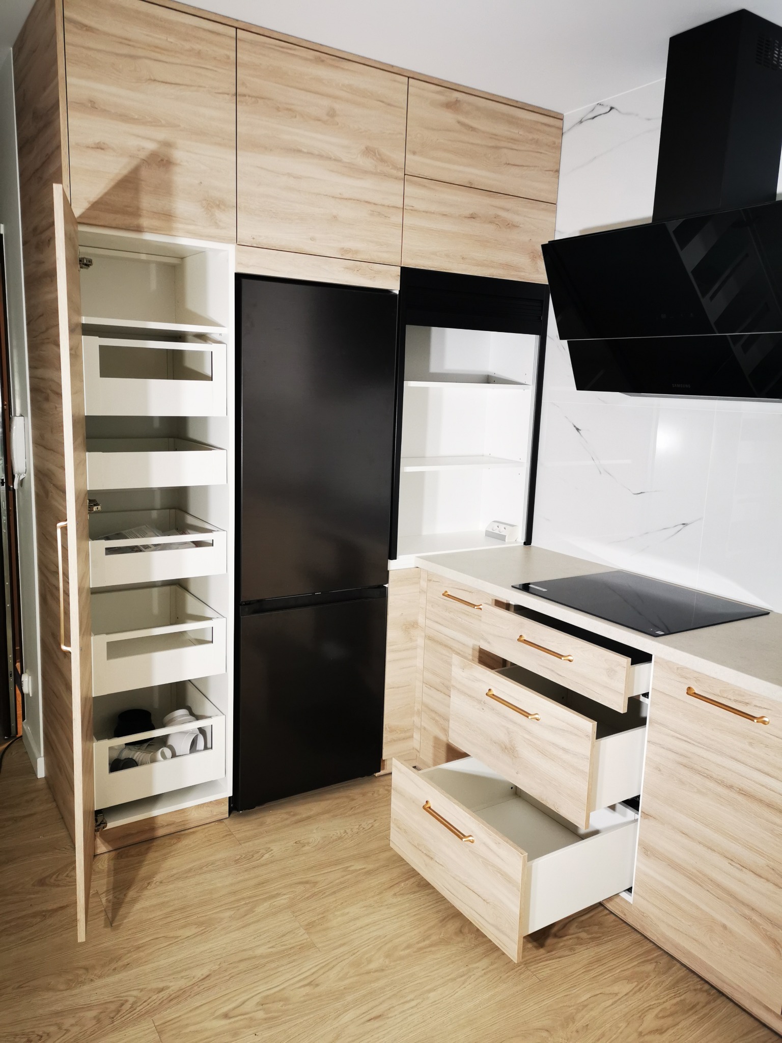 Jasna kuchnia z drewnopodobnymi frontami: lodówka w zabudowie, szafka cargo z wysuwanymi półkami, otwarte szuflady z podłużnymi uchwytami.