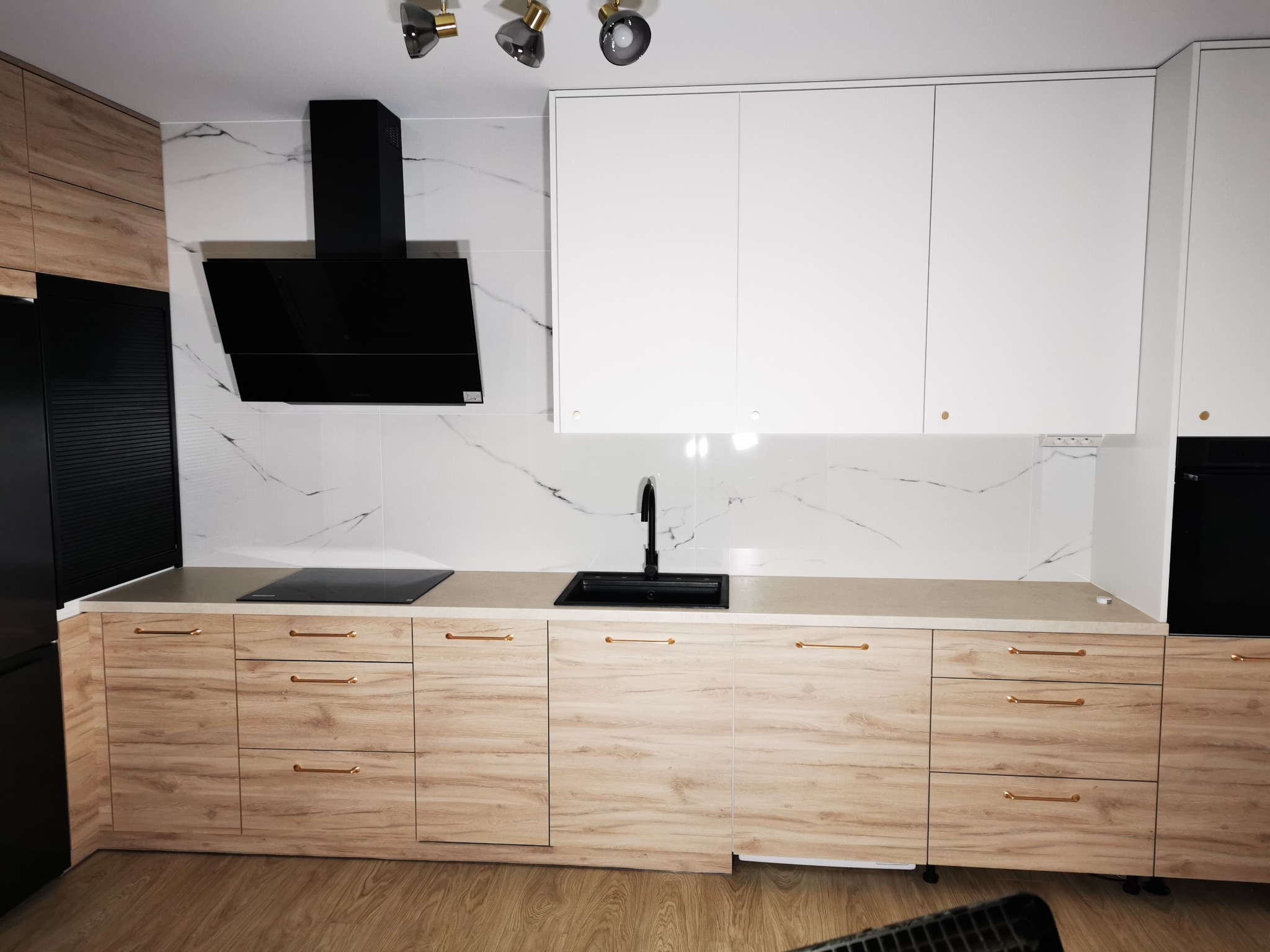Nowoczesna kuchnia z frontami w odcieniach drewna i białymi szafkami wiszącymi, czarny okap i armatura, marmurowe płytki na ścianie, oświetlenie punktowe.