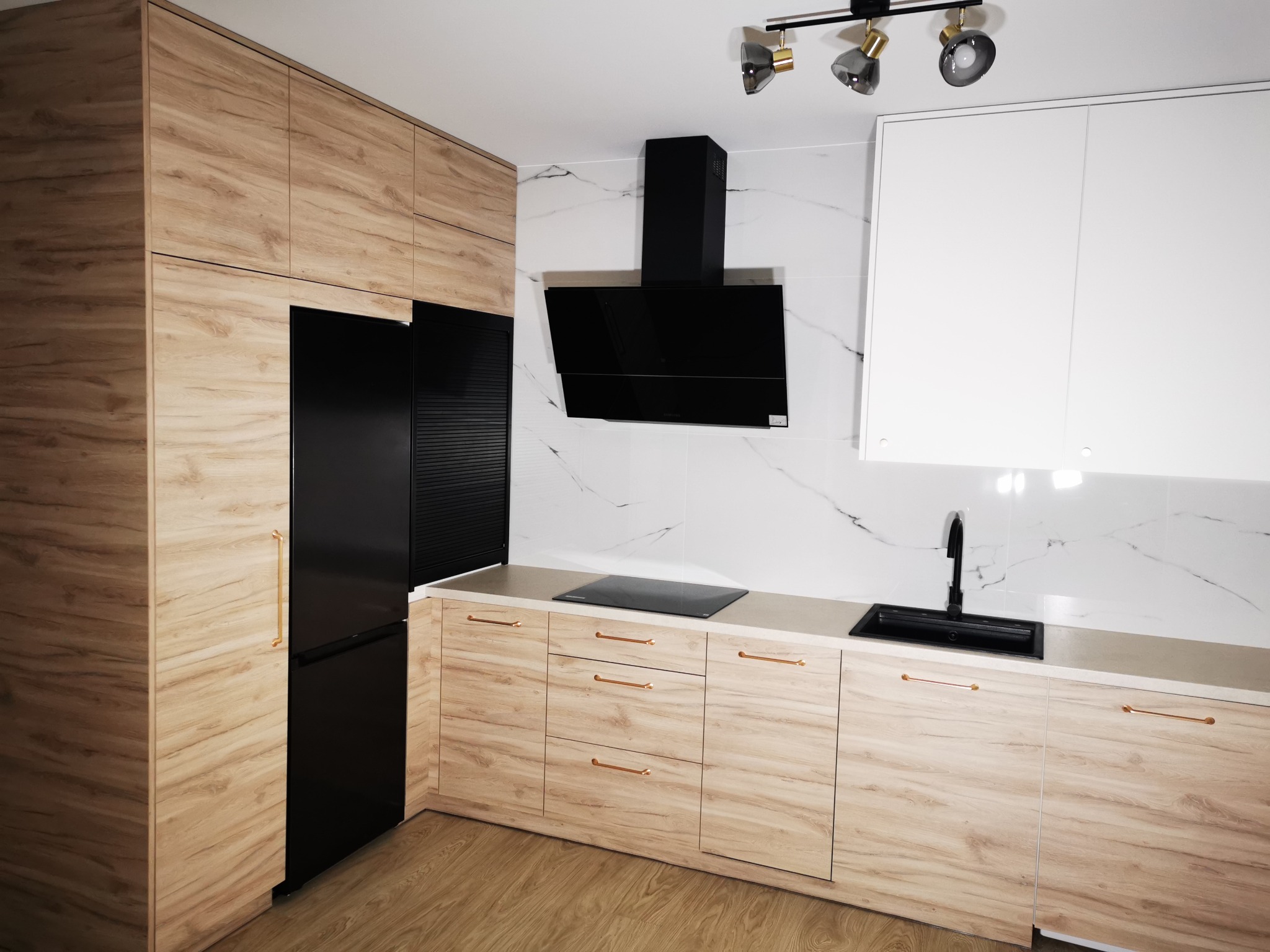 Nowoczesna kuchnia w Paterek z drewnianymi frontami, czarnym sprzętem AGD i marmurowym backsplashiem. Minimalistyczny design, złote uchwyty.