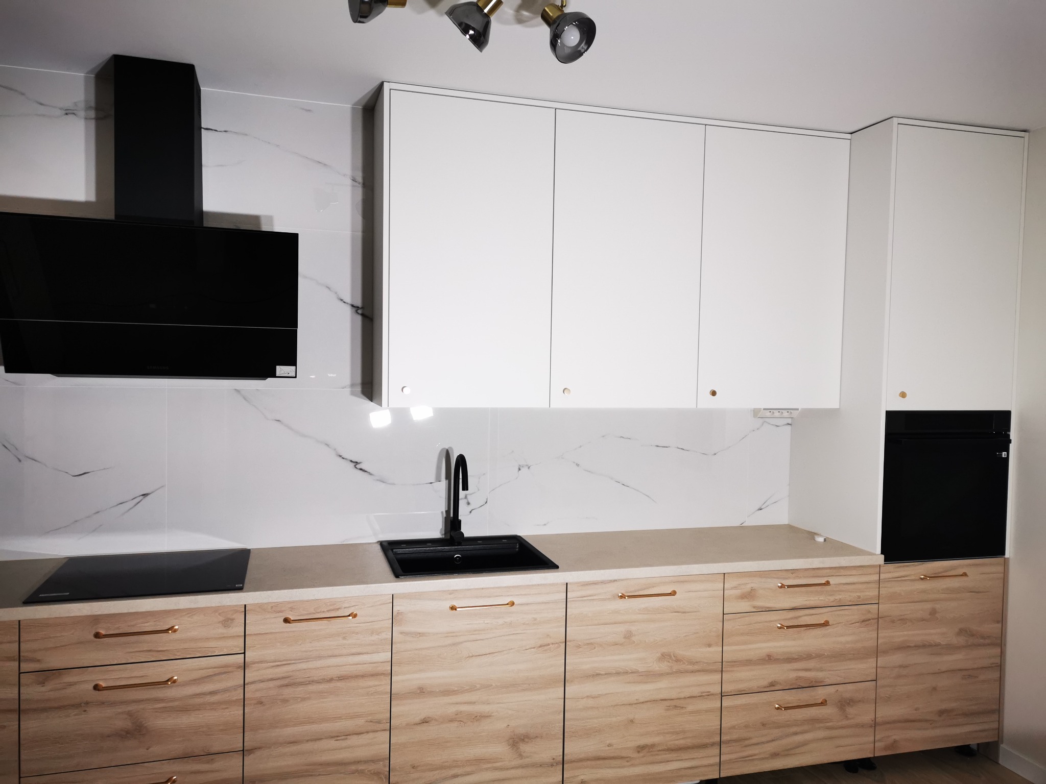 Nowoczesna kuchnia z białymi szafkami wiszącymi i drewnopodobnymi dolnymi. Czarny okap i zlew kontrastują z marmurowym tłem. Oświetlenie punktowe.