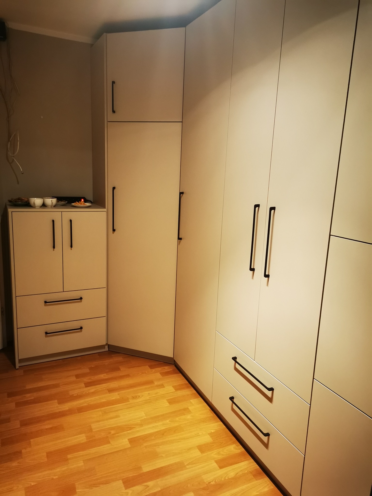 Jasna szafa narożna z czarnymi uchwytami na drewnianej podłodze. Widoczne drzwi i szuflady. Na szafce talerze i filiżanki.