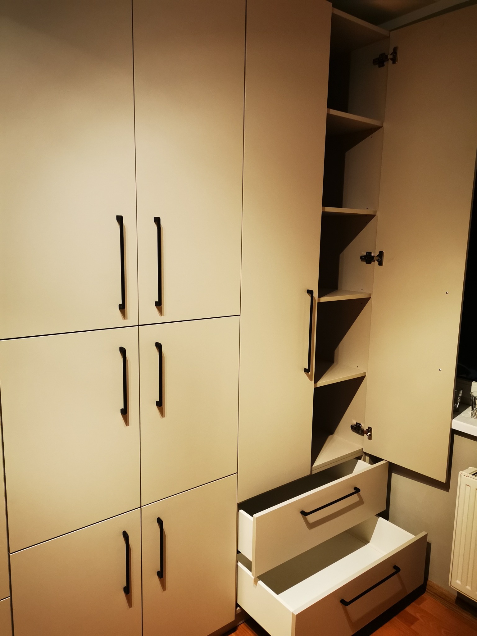 Jasna szafa wnękowa z czarnymi uchwytami. Jedna szafka otwarta, ukazująca półki, poniżej dwie wysunięte szuflady. Widoczne zawiasy i wnętrze szafy.