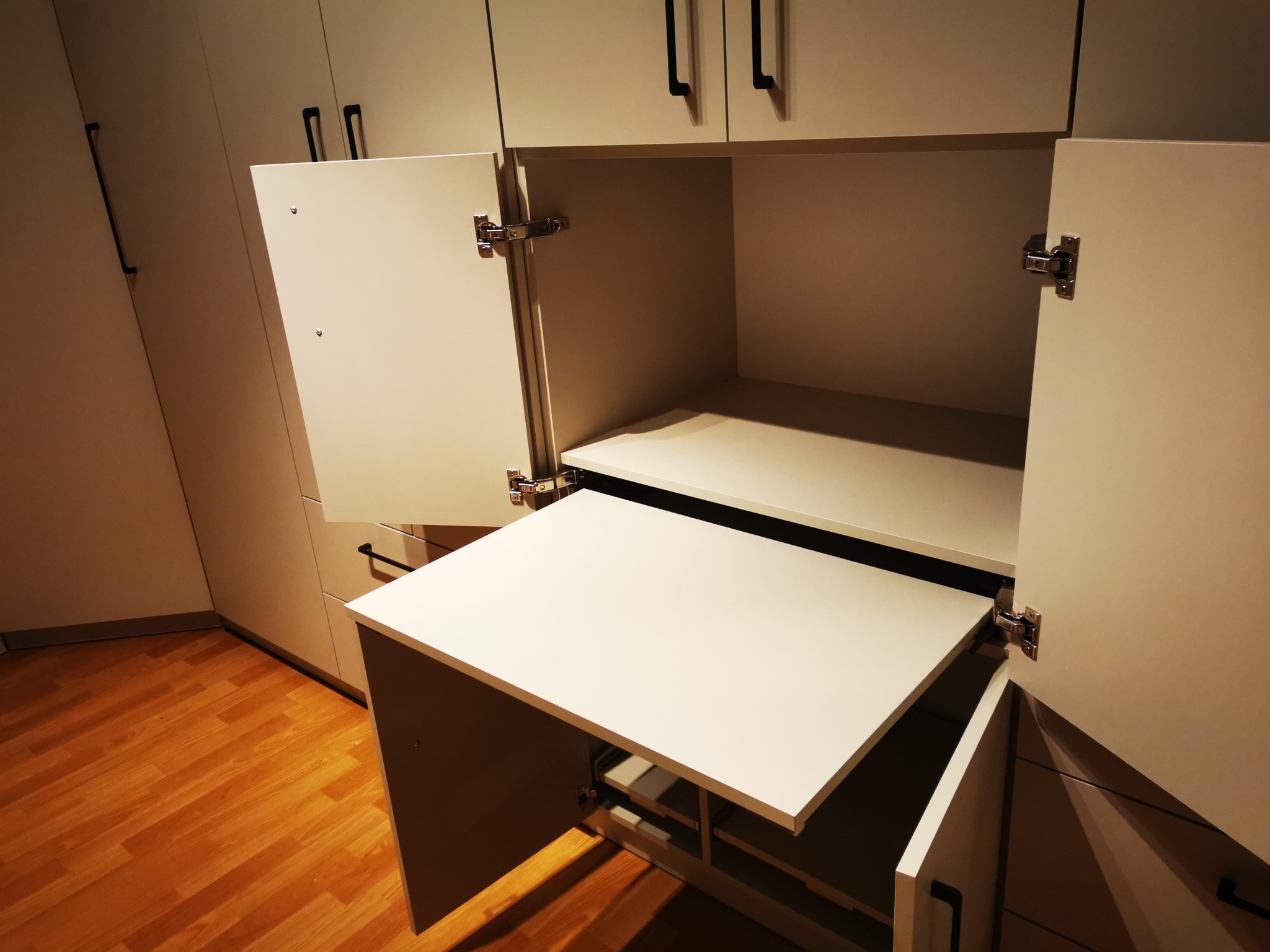 Biała szafa na wymiar z rozkładanym blatem, otwarte drzwiczki ukazują mechanizm. Widoczne minimalistyczne uchwyty i drewniana podłoga.