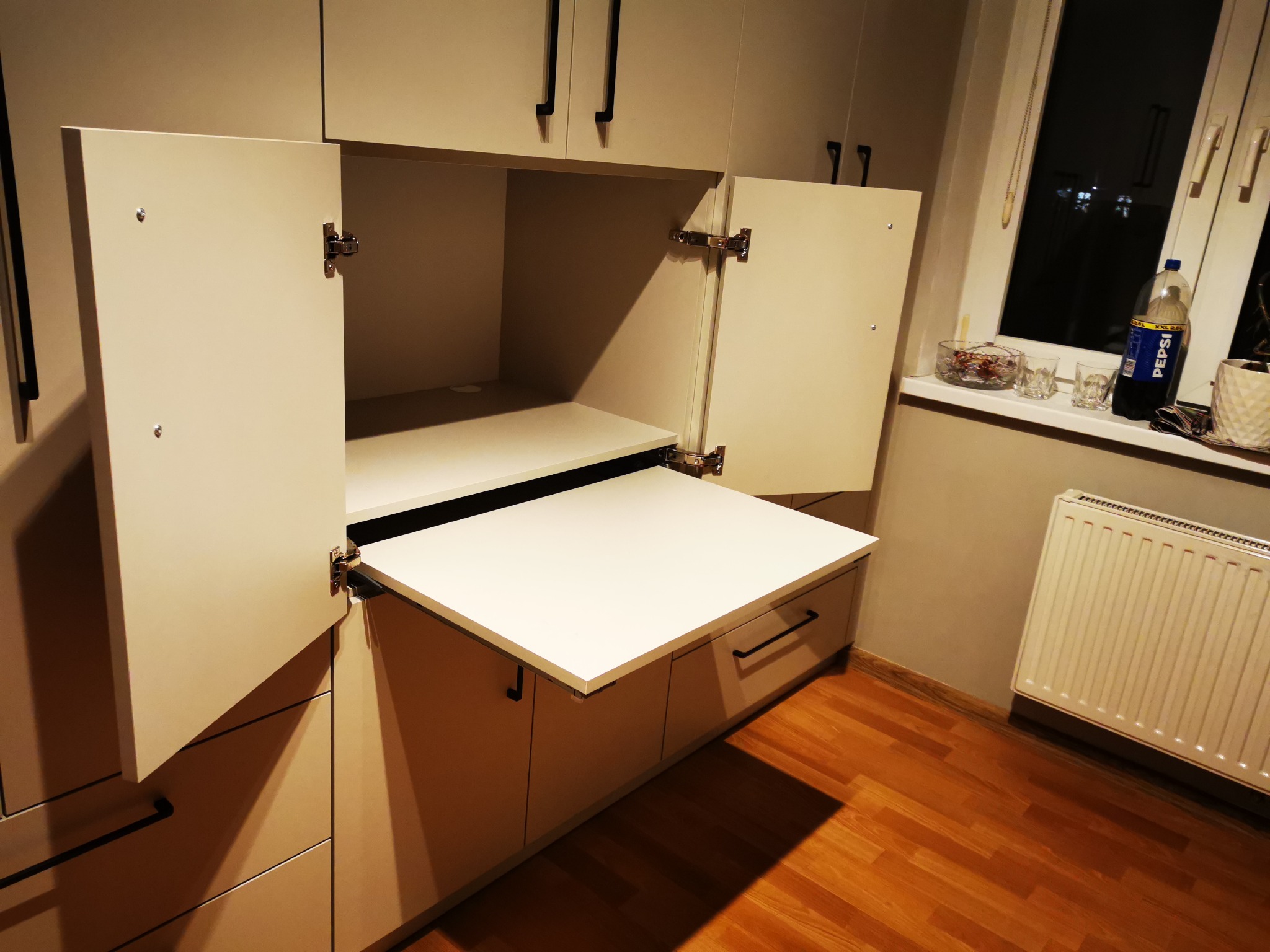 Beżowa szafa z wysuwanym, białym blatem. Otwarta szafka z wysuwanym blatem. Czarne uchwyty. Widoczne zawiasy. Na blacie przy oknie napój i naczynia.