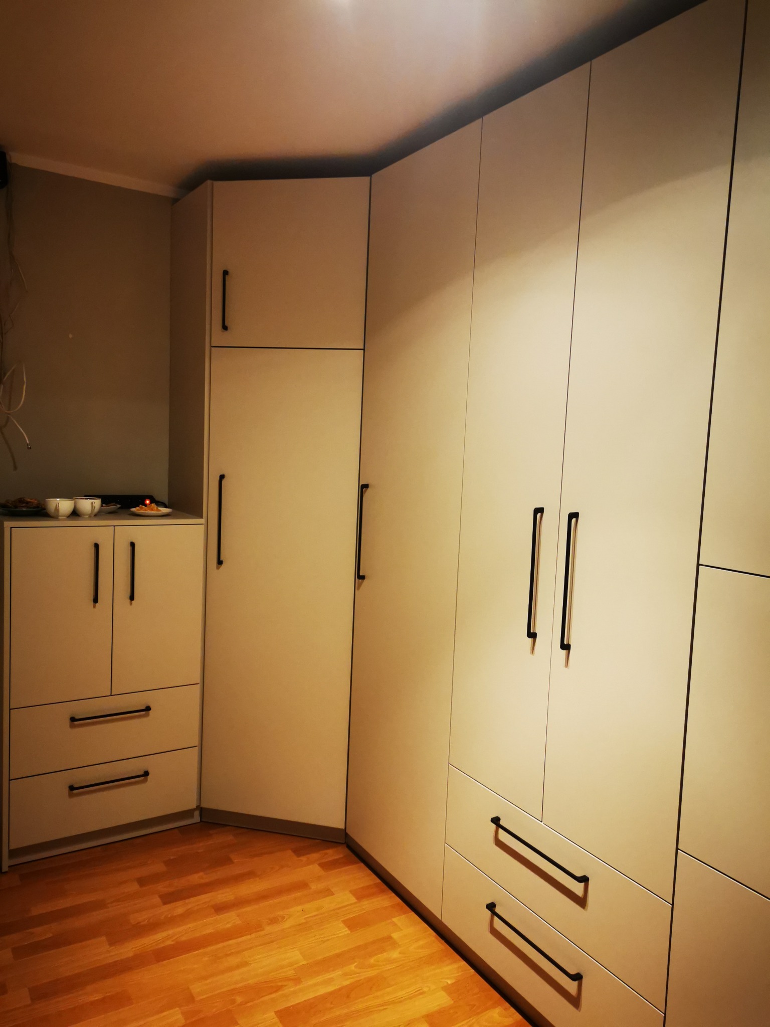 Jasna, narożna szafa z czarnymi, podłużnymi uchwytami, uzupełniona niską szafką z zastawą na blacie, na tle drewnianej podłogi.