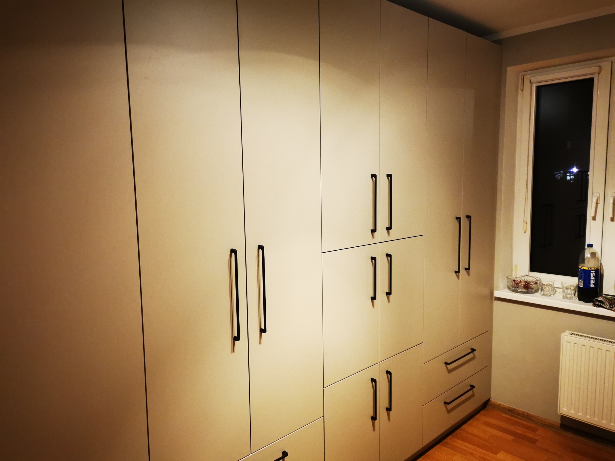 Jasna szafa wnękowa z czarnymi, podłużnymi uchwytami, ustawiona pod ścianą z oknem, na tle drewnianej podłogi.